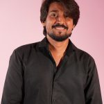 Portrait of a man in a black shirt smiling against a pink backdrop.