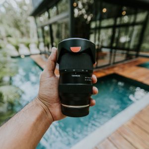 Close-up of a hand holding a Tamron camera lens by a poolside, outdoors.